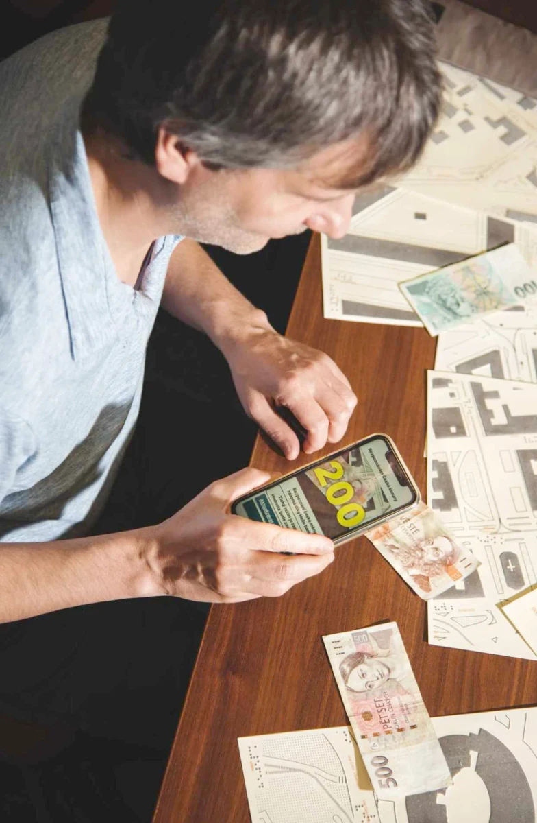رجل يجلس إلى مكتب عليه خرائط لمسية وأوراق نقدية يستخدم تطبيق Cash Reader الذي يعرض الرقم 200 بالأصفر.