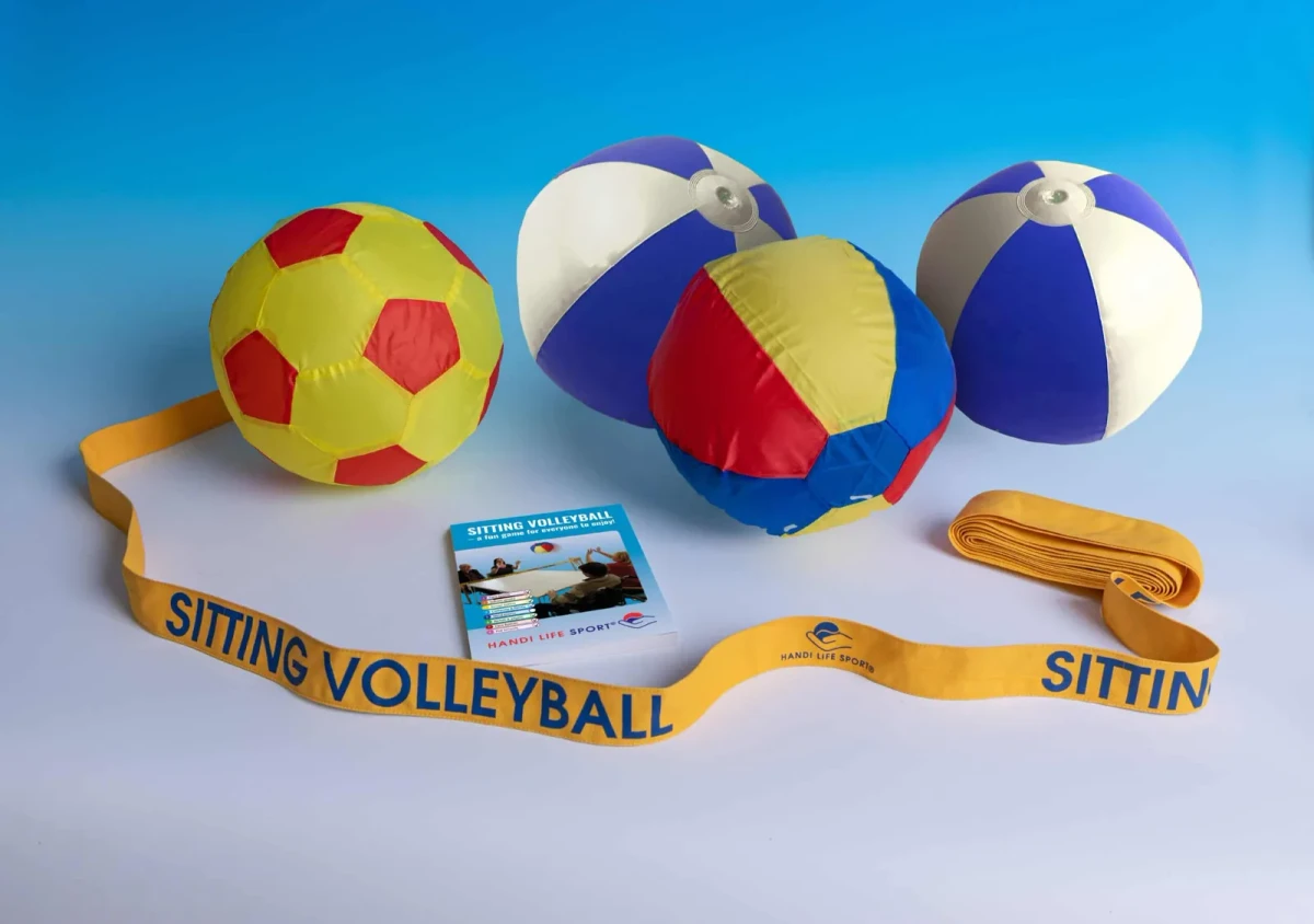 Ein Volleyball-Set: ein gelb-roter Ball, blau-weiße Strandbälle und ein gelbes Netzband mit der Aufschrift "Sitting Volleyball".