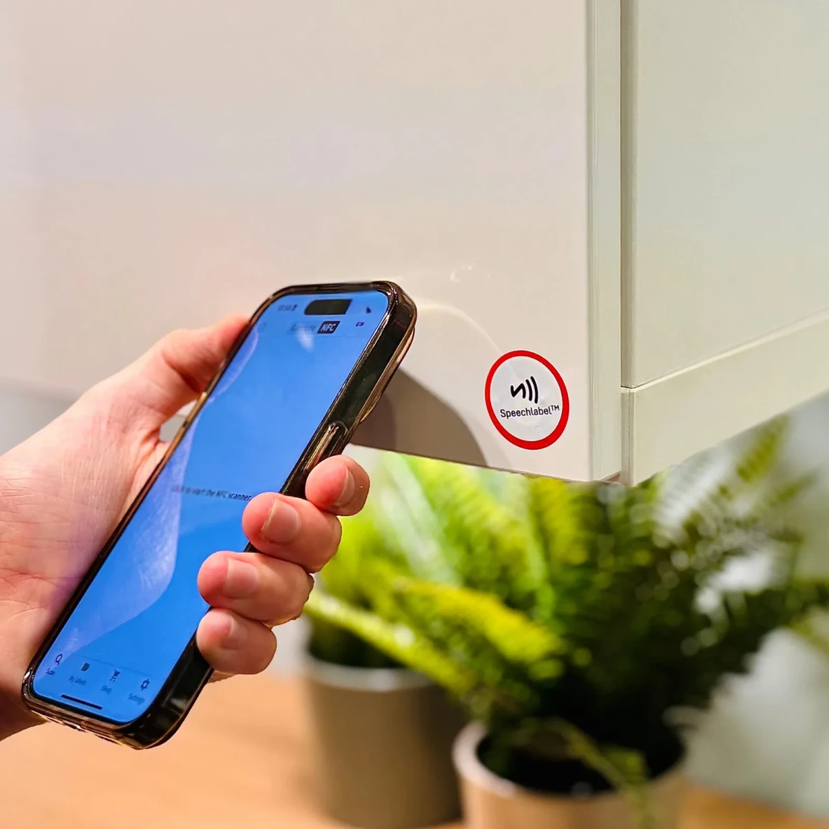 Eine Hand hält ein Smartphone an einen Speechlabel NFC-Aufkleber, der an der Seite eines weißen Schranks klebt, um ihn zu scannen. Darunter steht ein Topffarn.