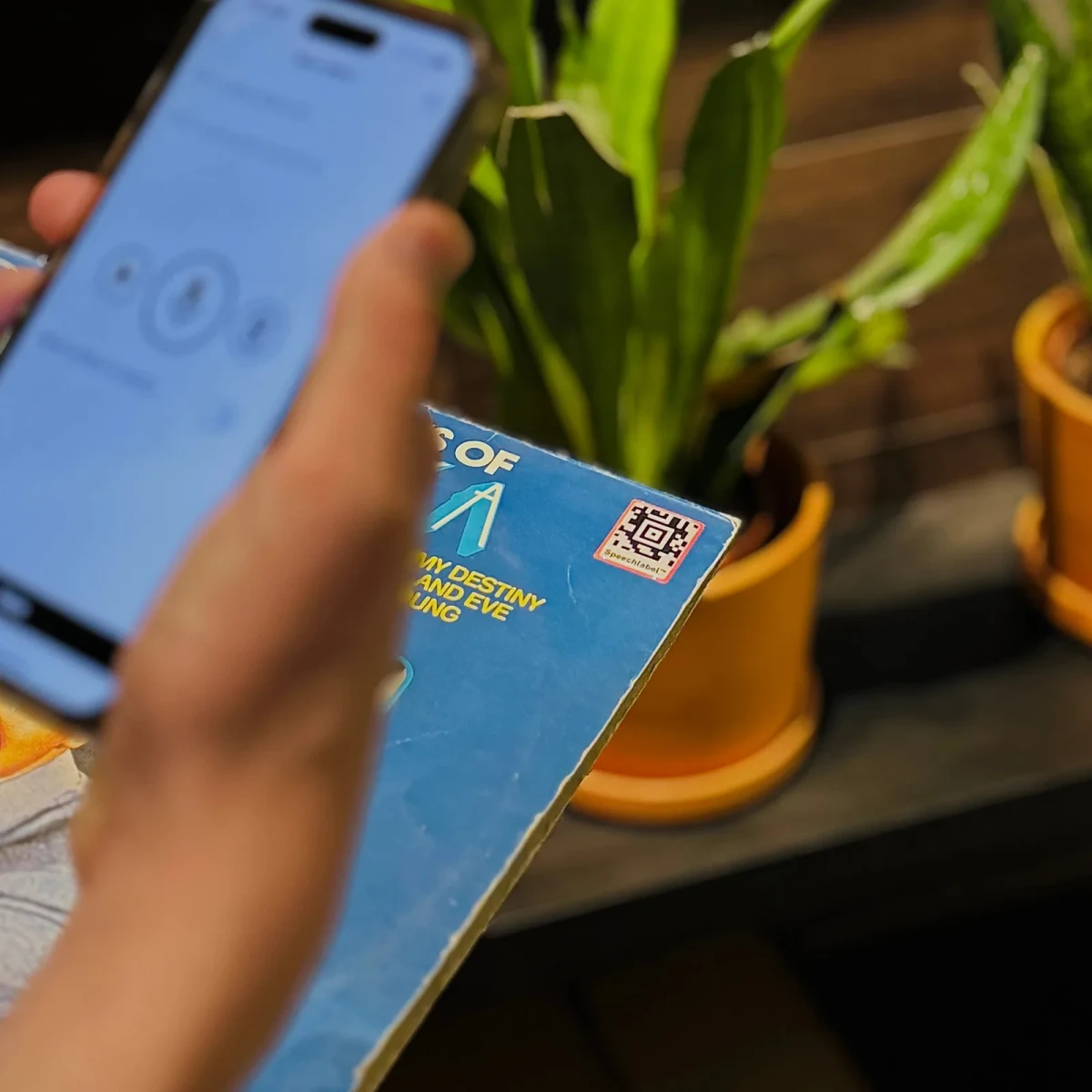 Eine Hand hält ein Smartphone an einen Speechlabel QR-Aufkleber, der auf der Ecke einer abgenutzten blauen Schallplattenhülle angebracht ist, um ihn zu scannen. Im Hintergrund stehen Topfpflanzen.