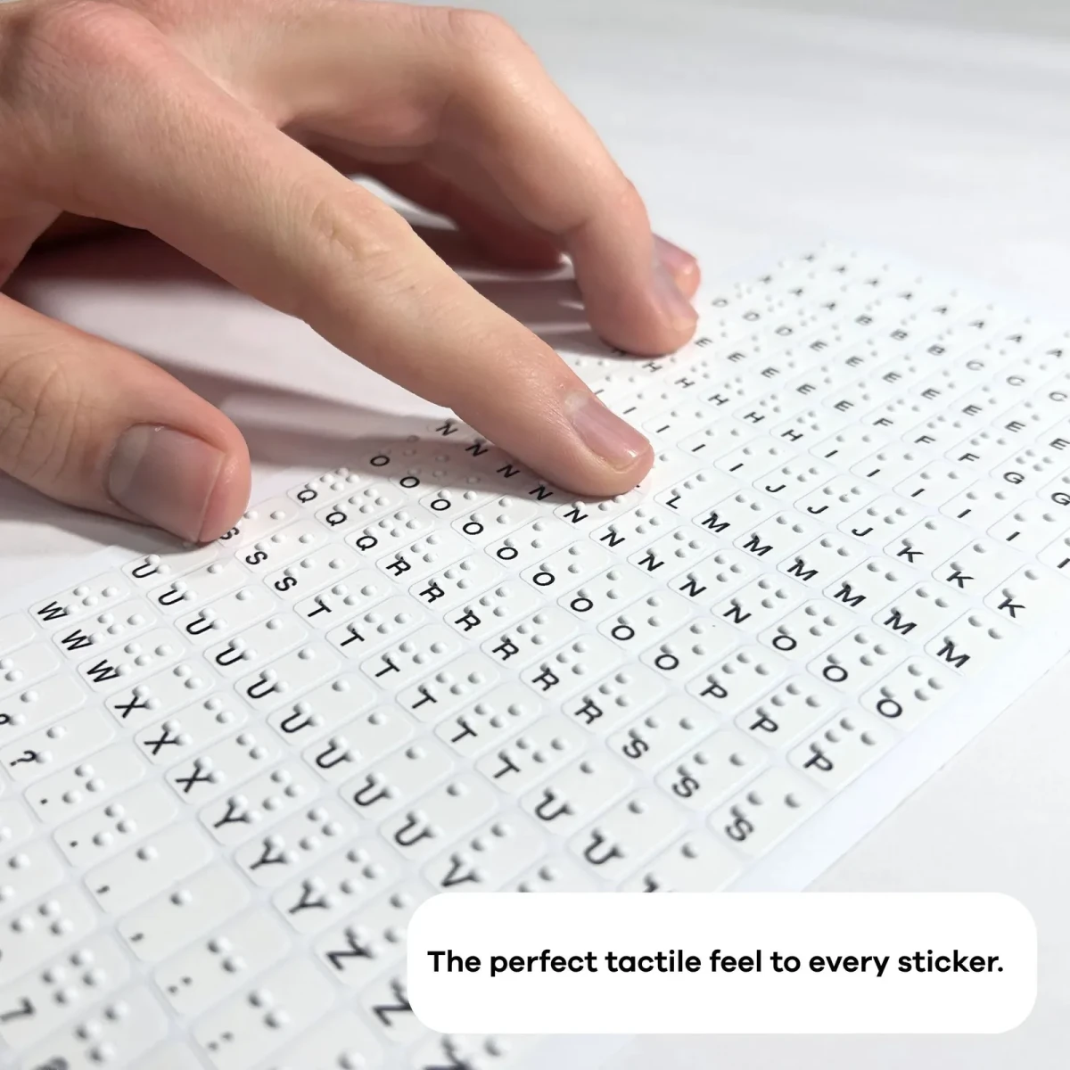 Eine Hand liegt auf dem flach liegenden kleinen Braille-Aufkleberbogen Hable Stactiles, der dichte Reihen kleiner Aufkleber zeigt, die jeweils erhabene Braillepunkte mit einem gedruckten Buchstaben kombinieren. Eine Bildunterschrift lautet: Das perfekte taktile Gefühl bei jedem Aufkleber.