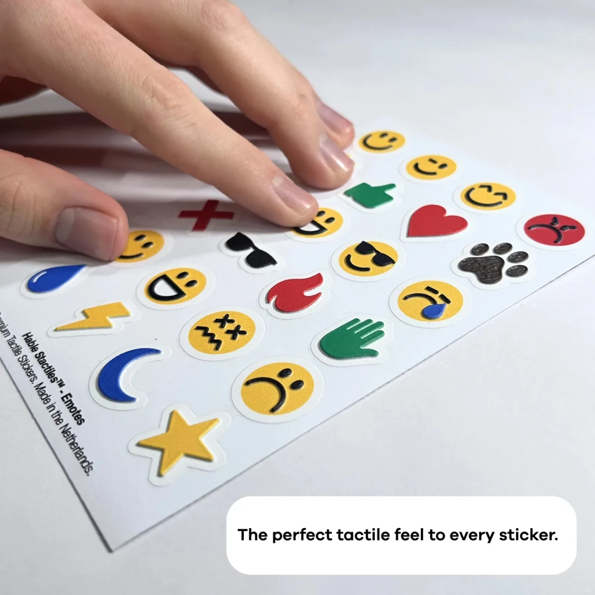 Una mano appoggiata sul foglio Hable Stactiles Emojis disteso, che mostra faccine sorridenti in rilievo, cuori, una fiamma, una stella e altre forme di emoji. Una didascalia recita: La perfetta sensazione tattile per ogni adesivo.