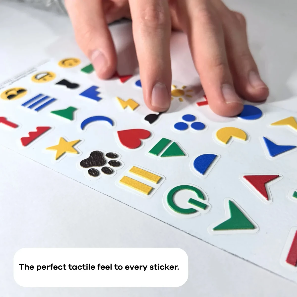 Una mano appoggiata sul foglio di Hable Stactiles Icons disteso, che mostra le varie icone in rilievo come una stella, un'impronta di zampa, un cuore, un sole e delle frecce. Una didascalia recita: La sensazione tattile perfetta per ogni adesivo.