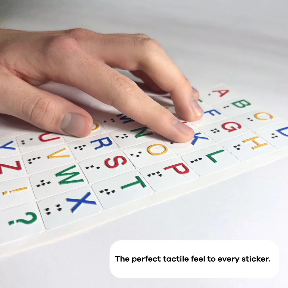 Una mano appoggiata sul foglio Hable Stactiles Letters disteso, che mostra lettere colorate in rilievo abbinate a punti Braille. Una didascalia recita: La perfetta sensazione tattile per ogni adesivo.
