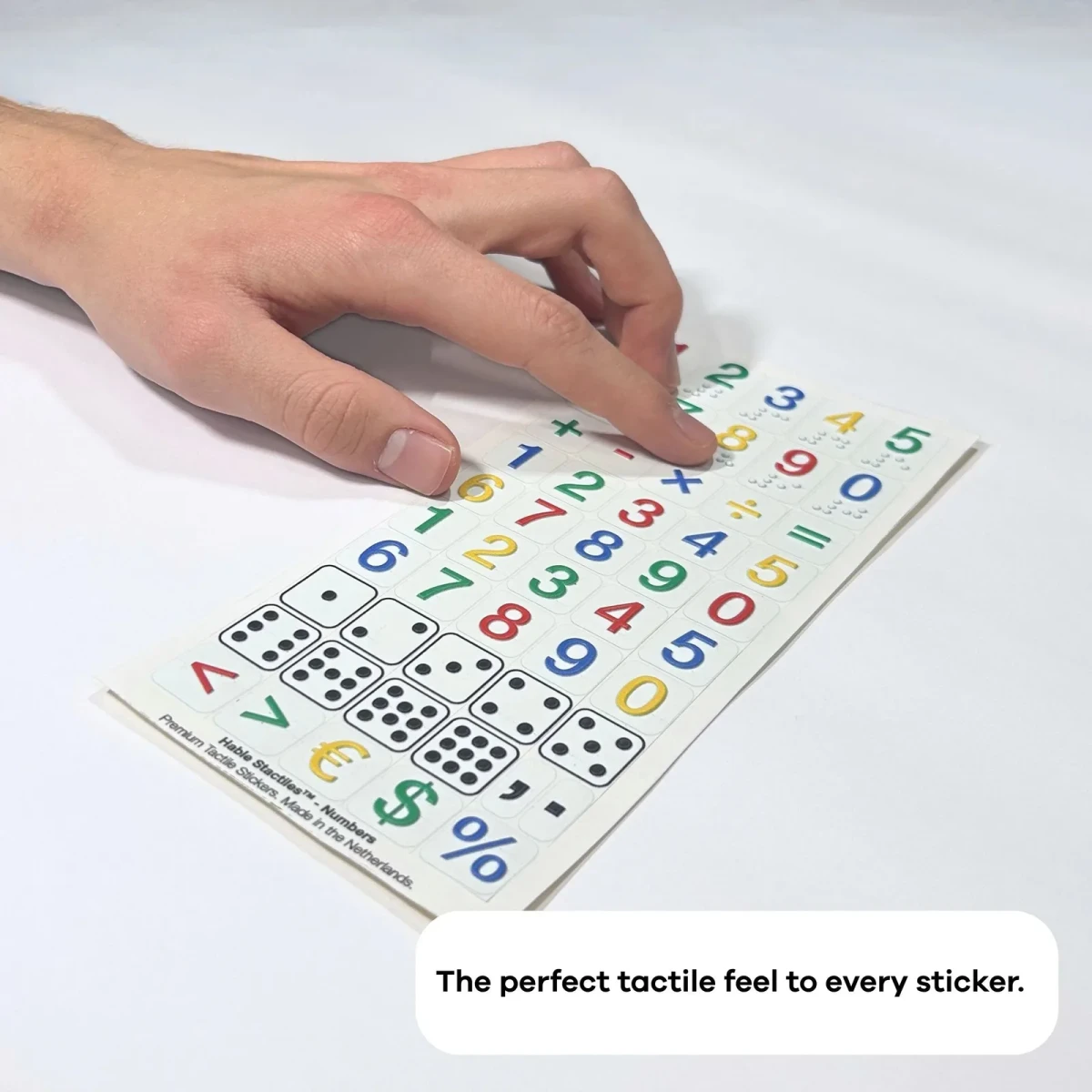 Eine Hand liegt auf dem flach liegenden Hable Stactiles Zahlenblatt, das erhabene farbige Ziffern, mathematische Symbole, Würfelmuster und Währungszeichen zeigt. Eine Bildunterschrift lautet: Die perfekte taktile Haptik für jeden Aufkleber.