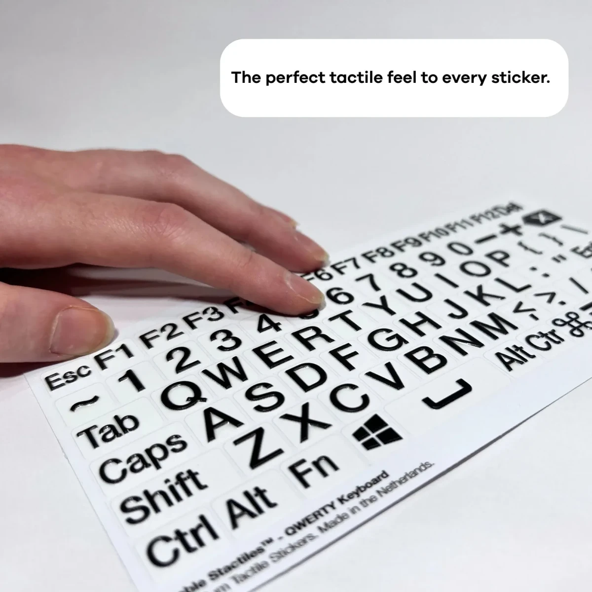 يد تستقر على ورقة Hable Stactiles بتخطيط QWERTY الموضوعة بشكل مسطح، تُظهر الحروف والأرقام ومفاتيح الوظائف البارزة باللون الأسود على خلفية بيضاء. يظهر تعليق يقول 'الملمس اللمسي المثالي لكل ملصق'.