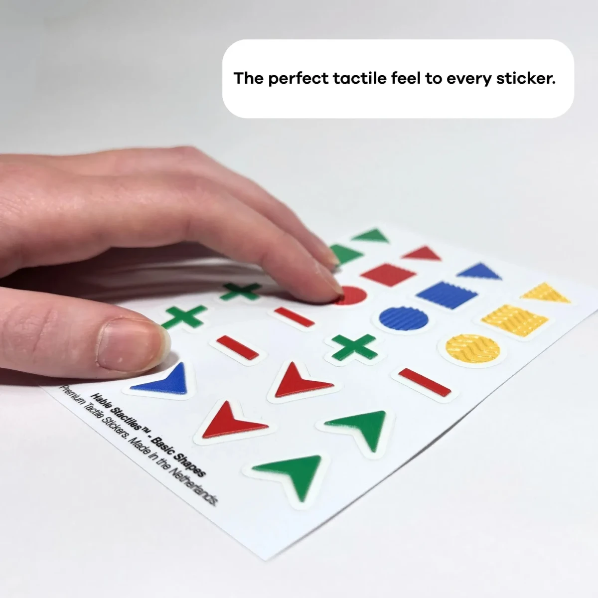 Una mano appoggiata sul foglio Hable Stactiles Forme Base disteso, che mostra triangoli, quadrati, cerchi e frecce in rilievo di diversi colori e texture. Una didascalia recita: 'La sensazione tattile perfetta per ogni adesivo'.