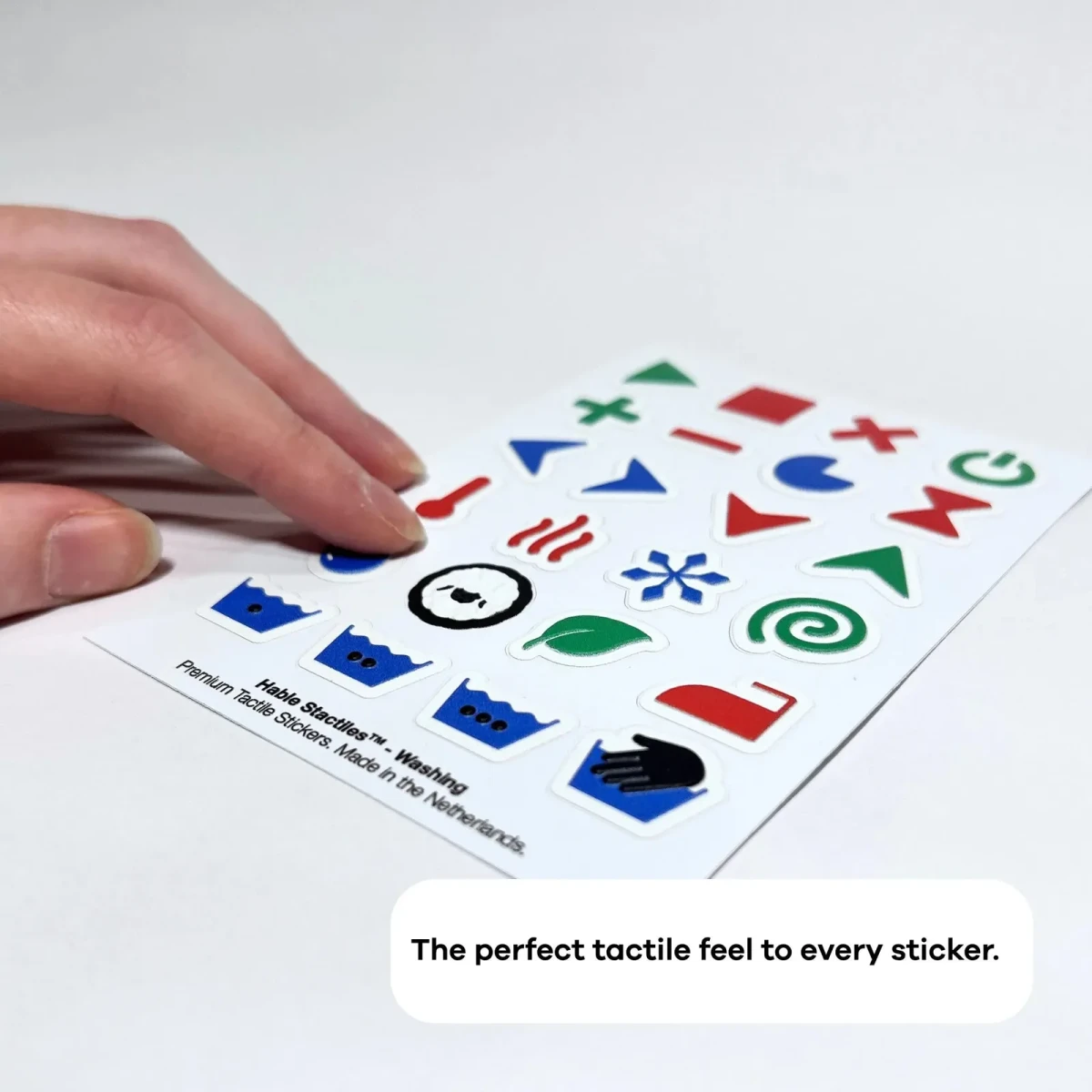 Eine Hand löst einen Aufkleber vom Hable Stactiles Waschbogen, der flach liegt und erhabene Wäschesymbole zeigt, darunter ein Thermometer, Hitzewellen, eine Schneeflocke, ein Blatt, ein Bügeleisen und Waschbottich-Symbole. Eine Bildunterschrift lautet: 'Das perfekte taktile Gefühl für jeden Aufkleber'.