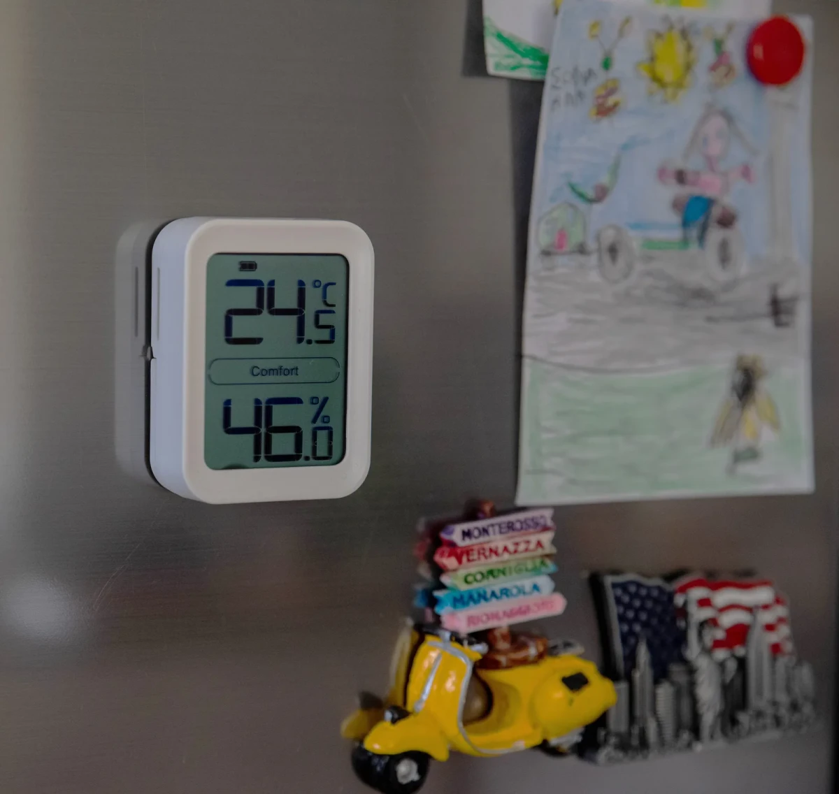 The TI-1 held by its magnet on a stainless steel fridge door among magnets and a child’s drawing